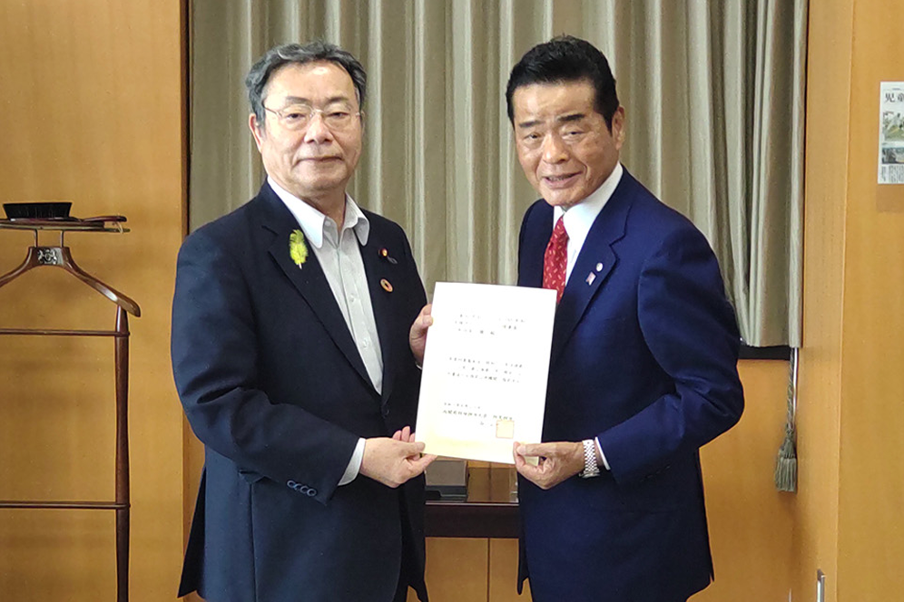 内閣府特命担当大臣の谷公一氏とAZ-COMネットワーク理事長の和佐見勝氏が指定通知書に関する文書を手に記念撮影している様子